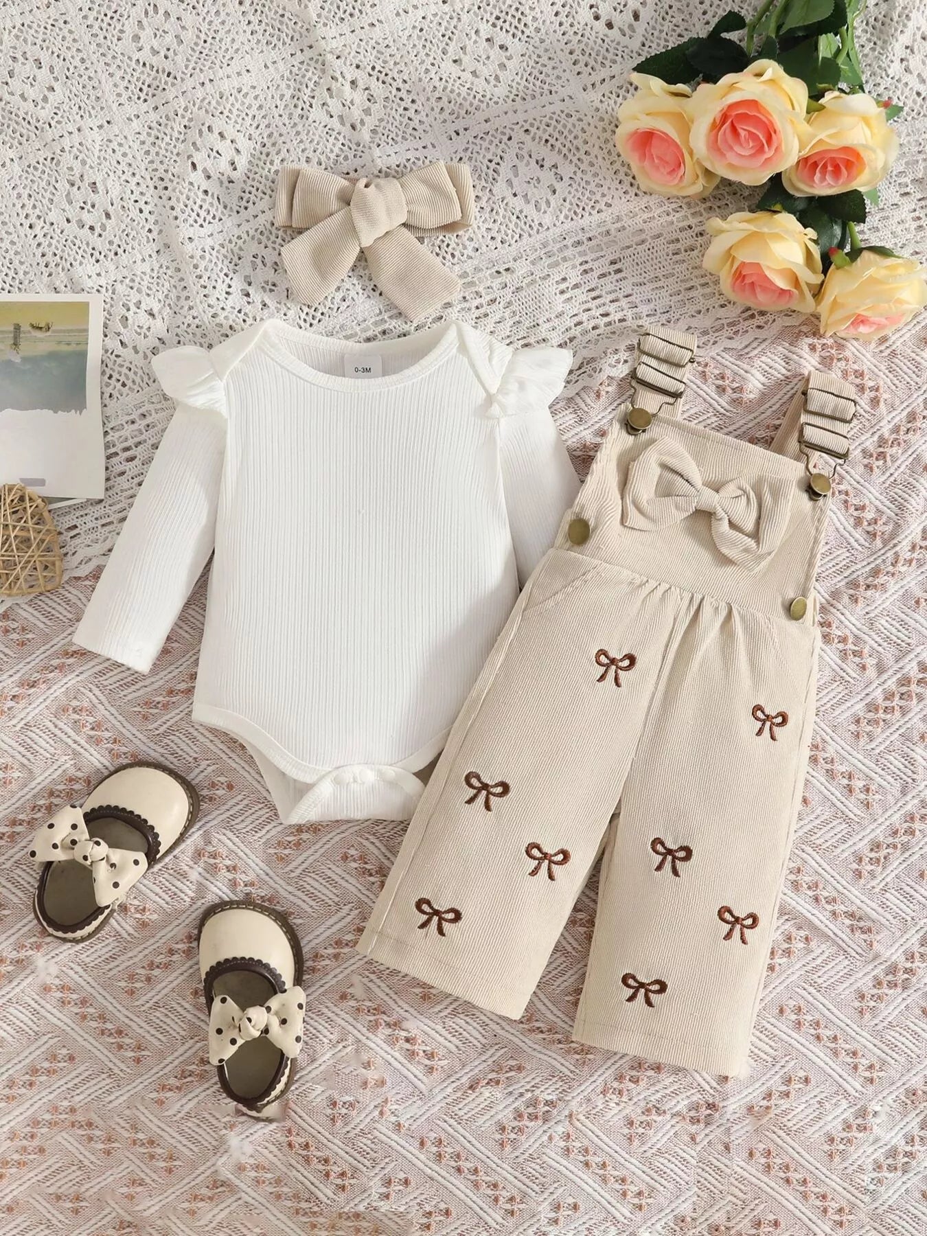 Baby girl three-piece outfit featuring a cream ribbed long-sleeve bodysuit with ruffled shoulders, apricot color bow-embroidered overalls and matching apricot bow headband.