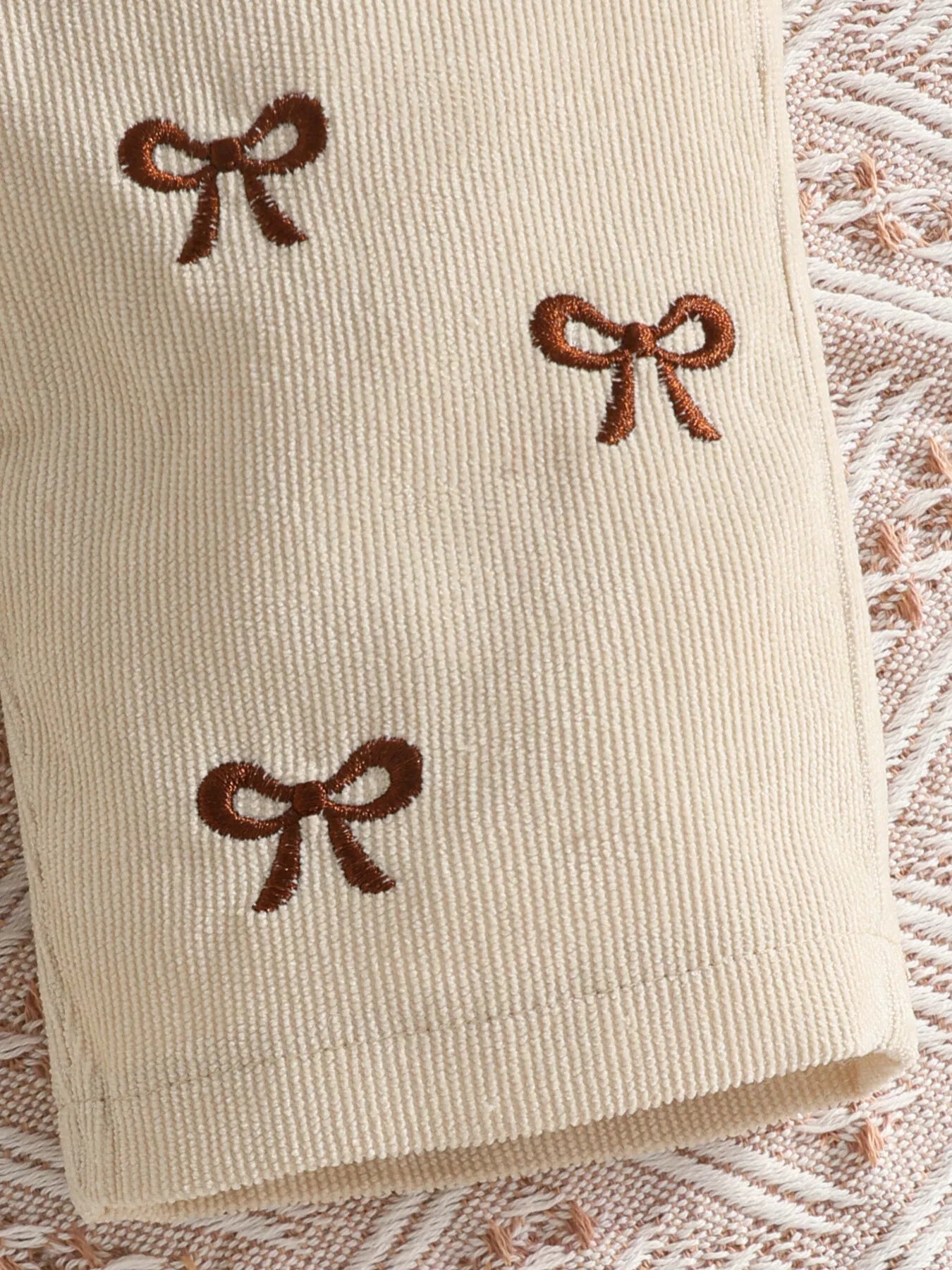 Baby girl three-piece outfit featuring a cream ribbed long-sleeve bodysuit with ruffled shoulders, apricot color bow-embroidered overalls and matching apricot bow headband.