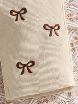 Baby girl three-piece outfit featuring a cream ribbed long-sleeve bodysuit with ruffled shoulders, apricot color bow-embroidered overalls and matching apricot bow headband.