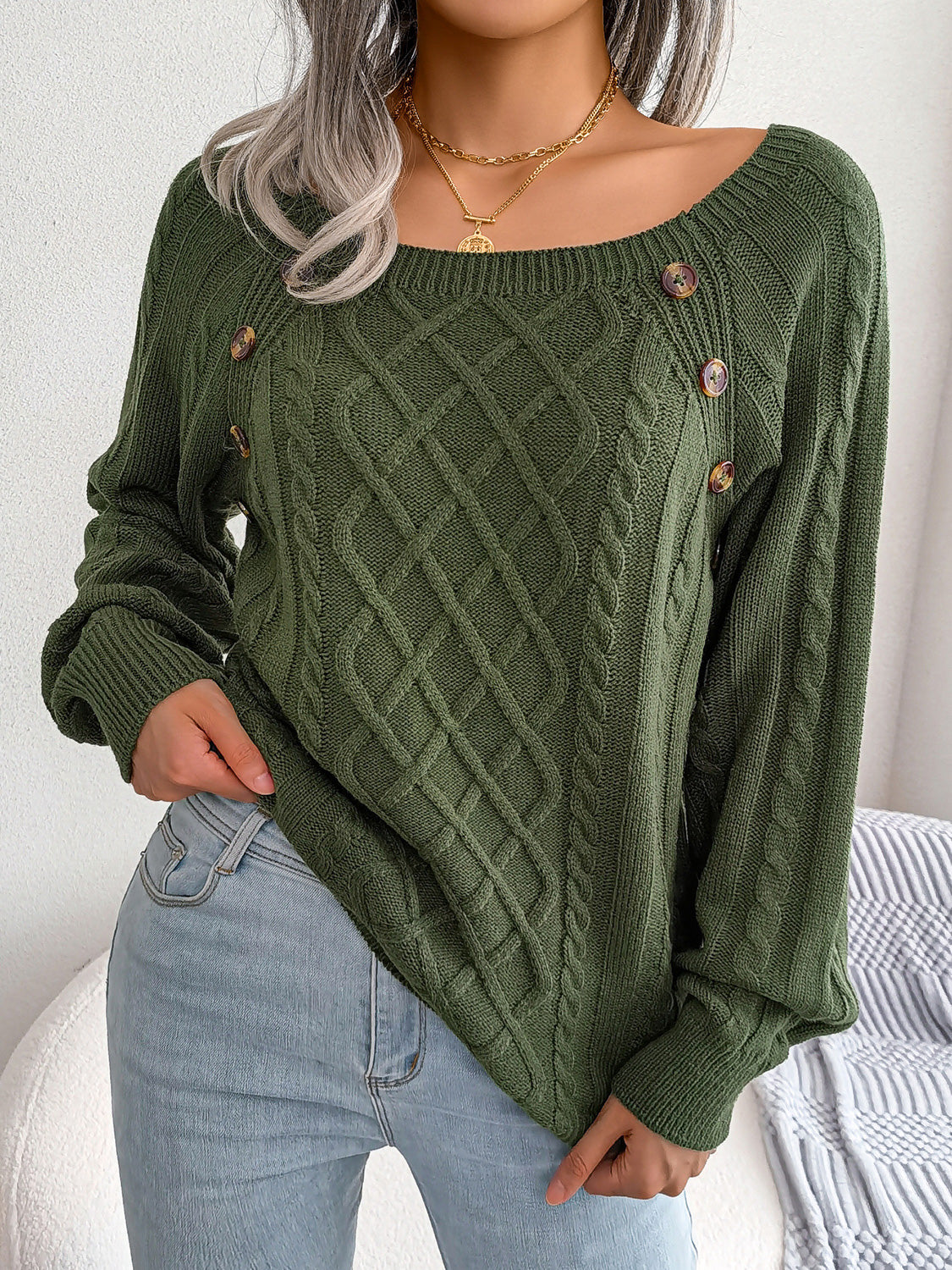 Woman wearing an army green cable-knit sweater with decorative buttons, paired with light-wash jeans, layered gold necklaces, and a white handbag. Perfect for cozy fall and winter outfits.