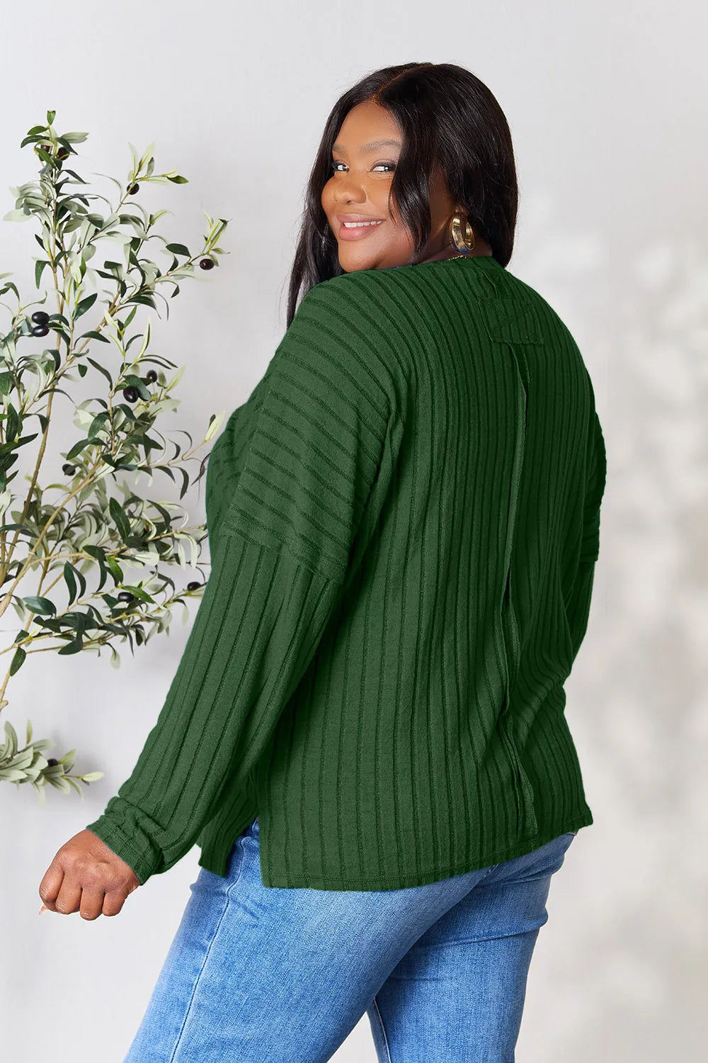 Woman wearing a dark green ribbed long sleeve knit top with a round neckline and side slit detail.