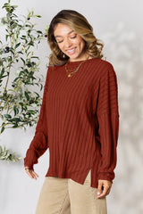 Woman wearing a rust ribbed long sleeve knit top with a round neckline and side slit detail.