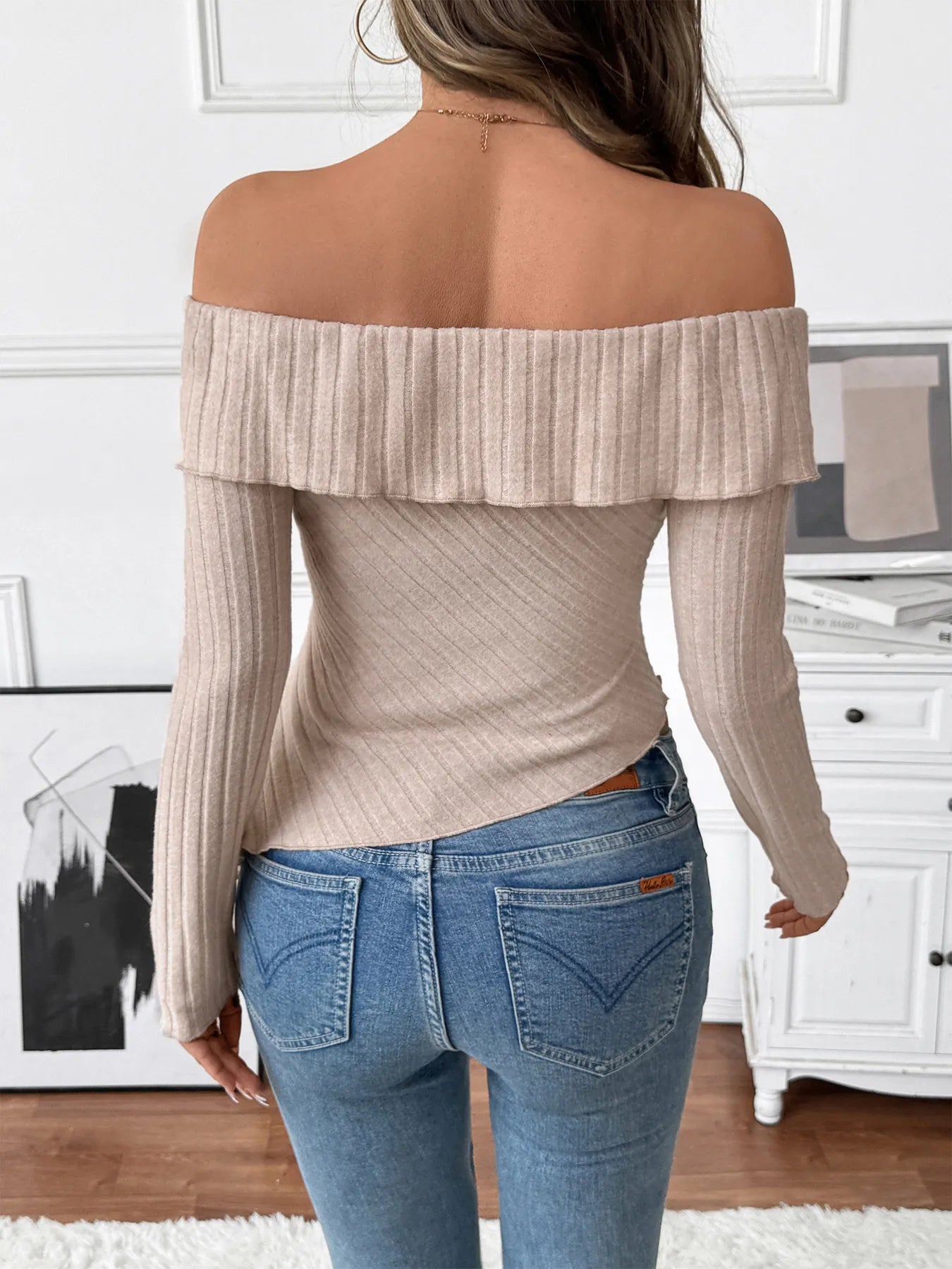 Woman wearing a beige asymmetrical off-shoulder ribbed knit top with twisted neckline and long sleeves paired with blue jeans.