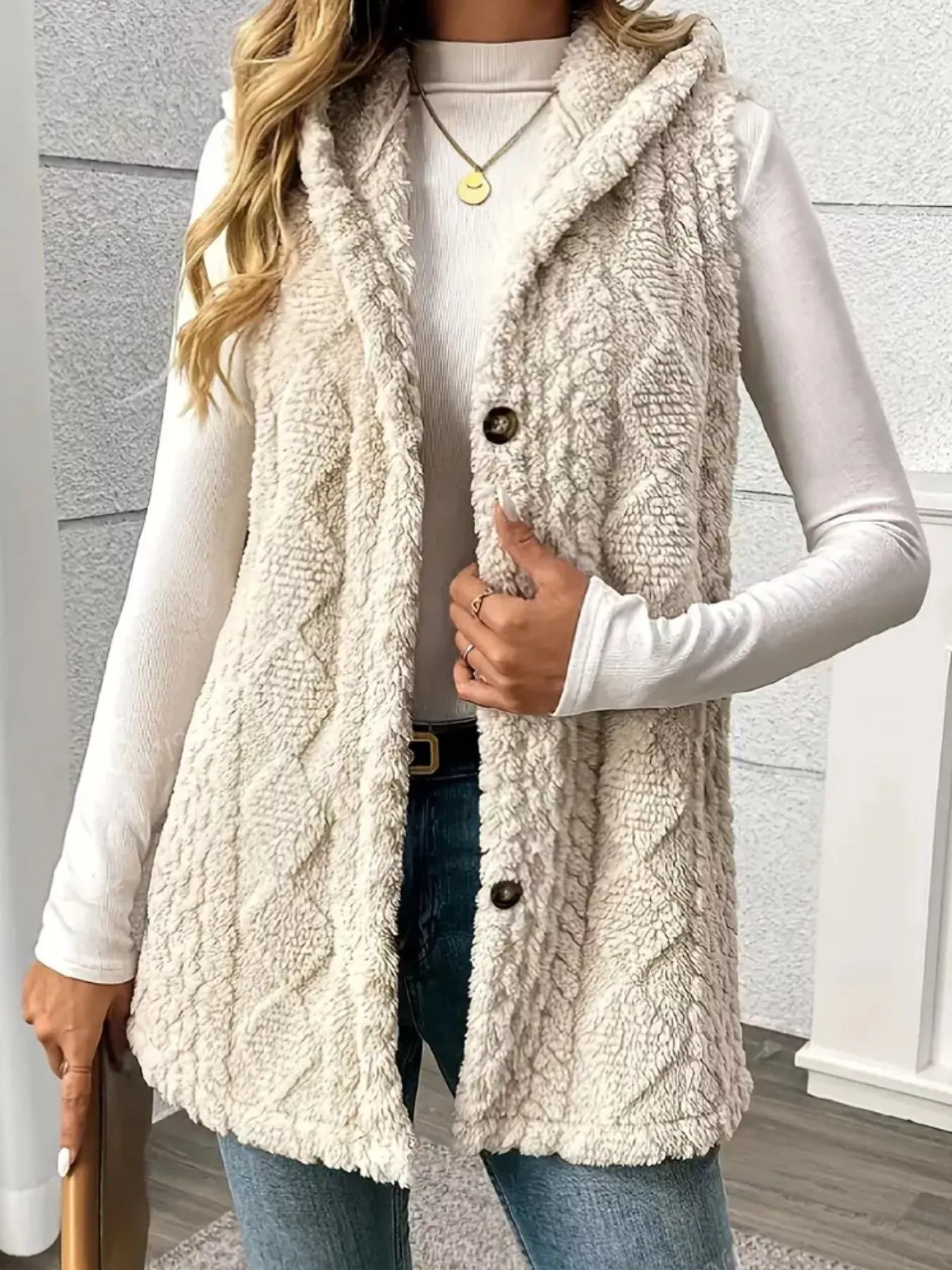 Woman wearing a beige hooded sherpa vest coat over a white long-sleeve top with blue jeans for a cozy fall and winter outfit.