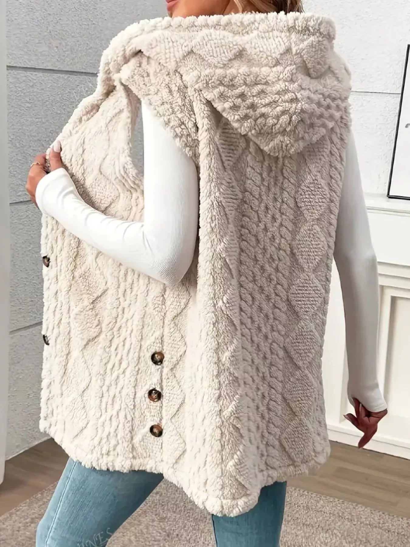 Woman wearing a beige hooded sherpa vest coat over a white long-sleeve top with blue jeans for a cozy fall and winter outfit.