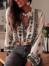 Woman wearing a beige and black vintage print long-sleeve blouse with dark denim jeans, a brown belt, and statement jewelry.
