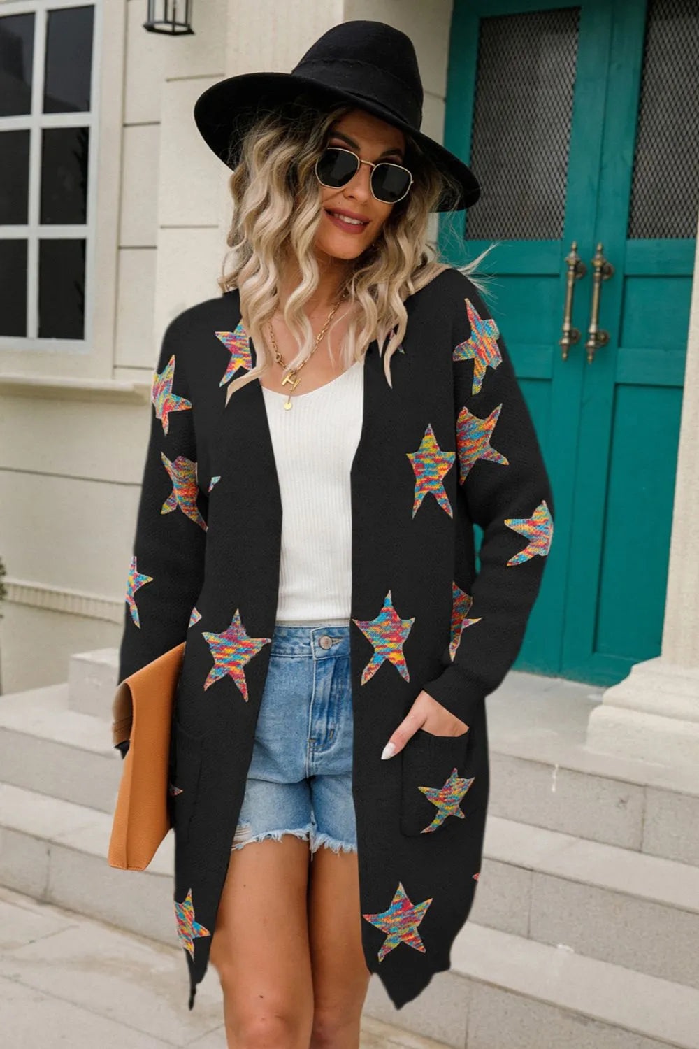 Woman wearing a black open-front cardigan with colorful star print, front pockets, denim shorts and black Western boots.