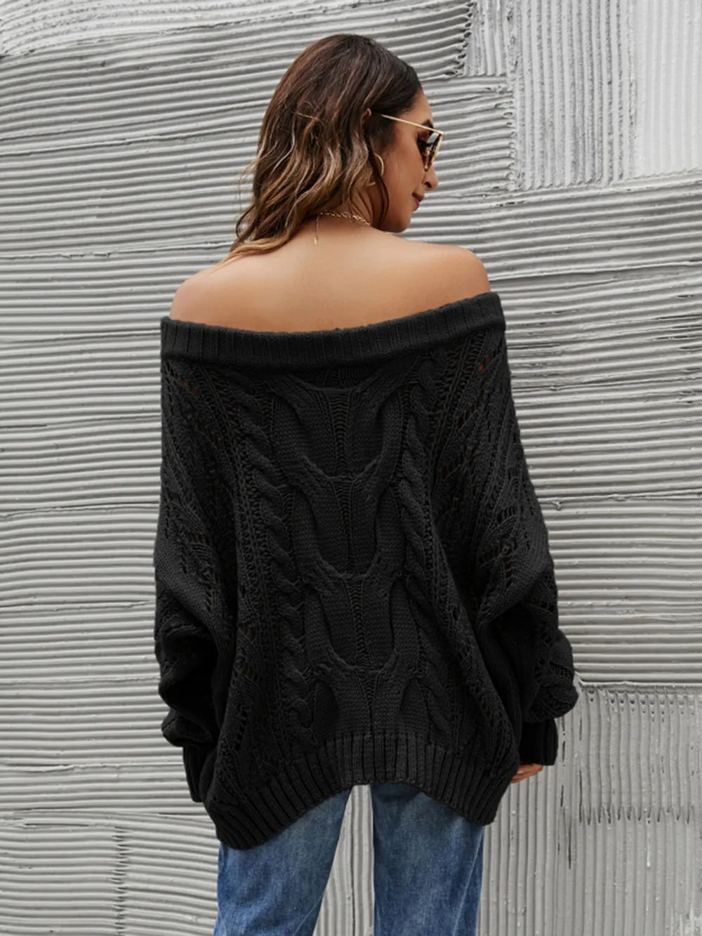 Woman wearing a black cable knit openwork off-shoulder sweater with distressed jeans and a wide-brim hat.