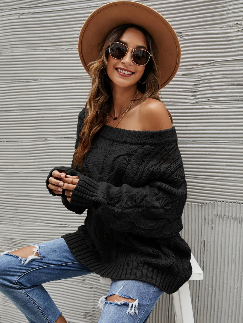 Woman wearing a black cable knit openwork off-shoulder sweater with distressed jeans and a wide-brim hat.