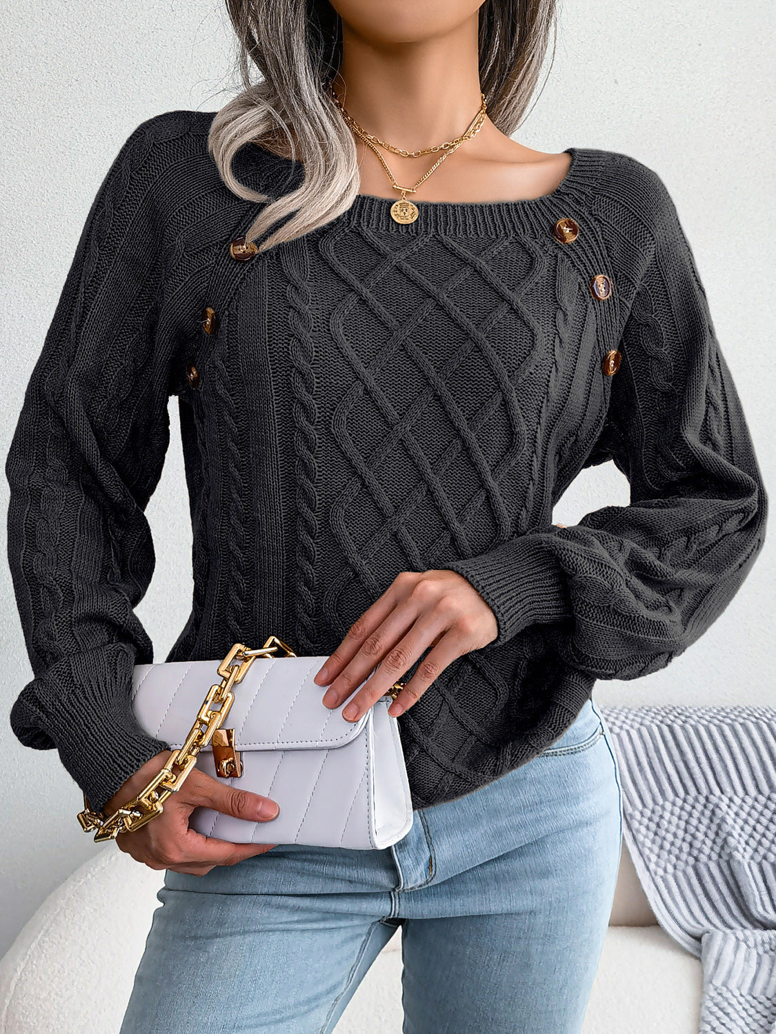 Woman wearing a black cable-knit sweater with decorative buttons, paired with light-wash jeans, layered gold necklaces, and a white handbag. Perfect for cozy fall and winter outfits.