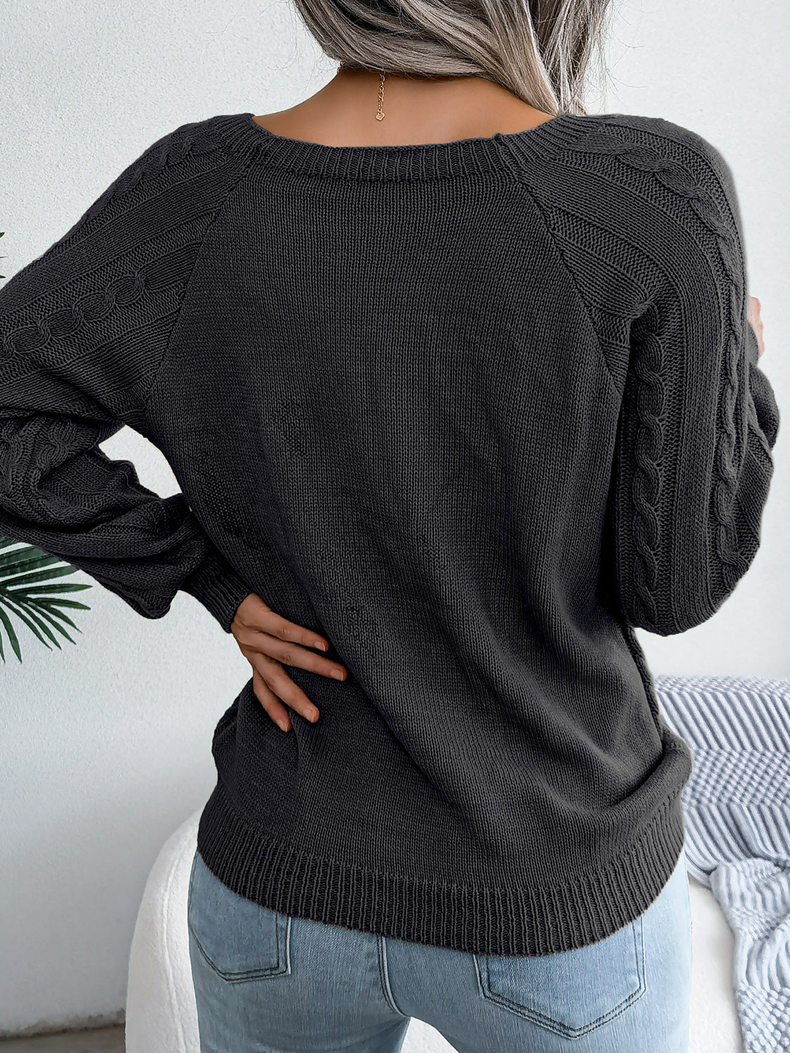 Woman wearing a black cable-knit sweater with decorative buttons, paired with light-wash jeans, layered gold necklaces, and a white handbag. Perfect for cozy fall and winter outfits.