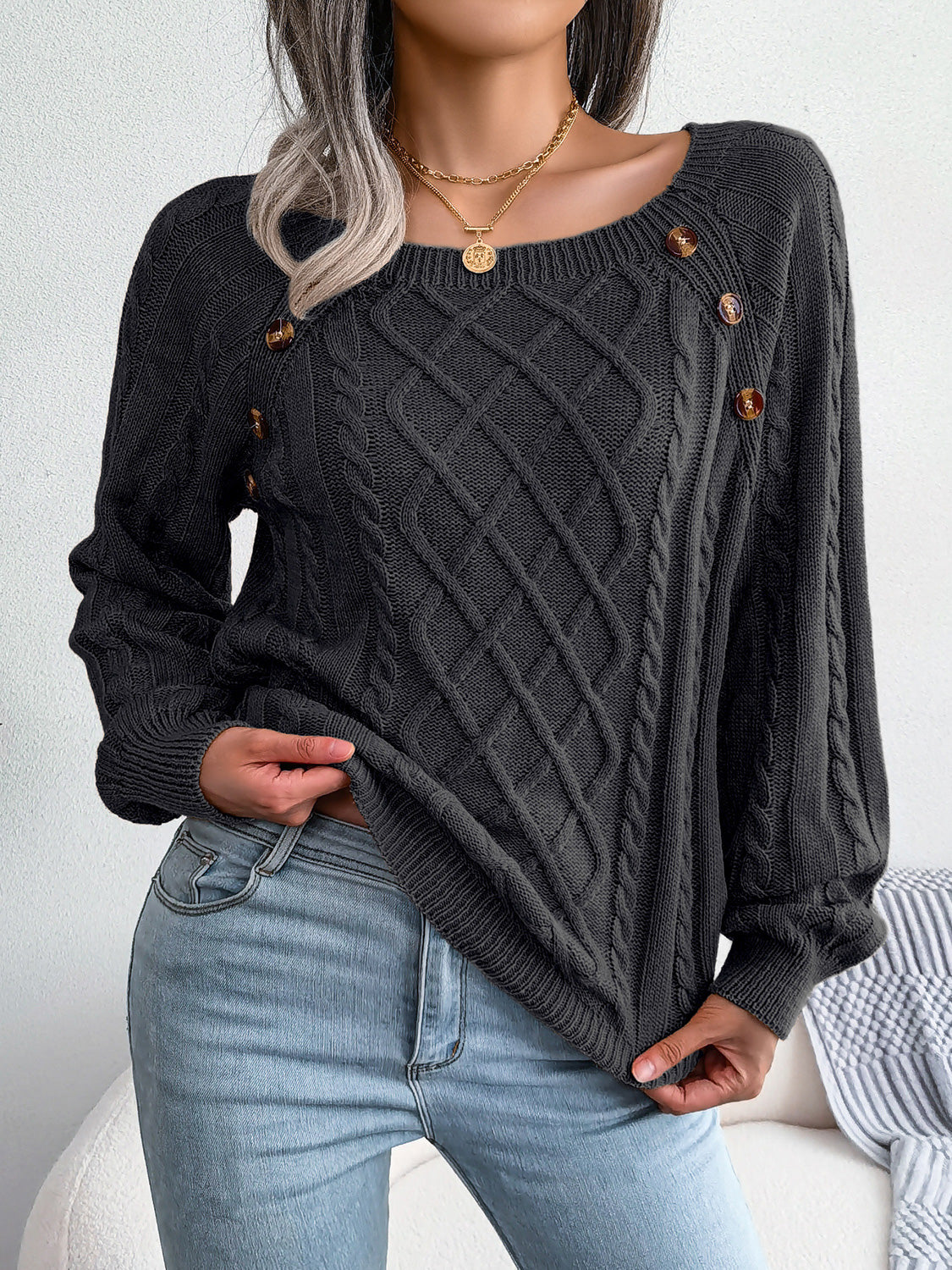 Woman wearing a black cable-knit sweater with decorative buttons, paired with light-wash jeans, layered gold necklaces, and a white handbag. Perfect for cozy fall and winter outfits.