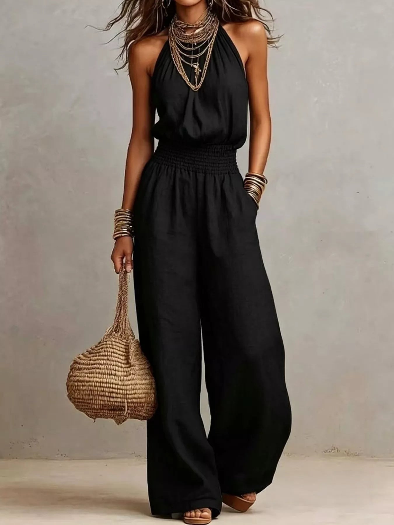 Woman wearing a black halter-neck wide-leg jumpsuit with a smocked waist, layered gold jewelry, and bracelets, styled for an elegant evening look.