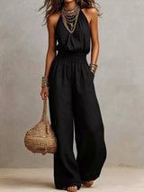 Woman wearing a black halter-neck wide-leg jumpsuit with a smocked waist, layered gold jewelry, and bracelets, styled for an elegant evening look.