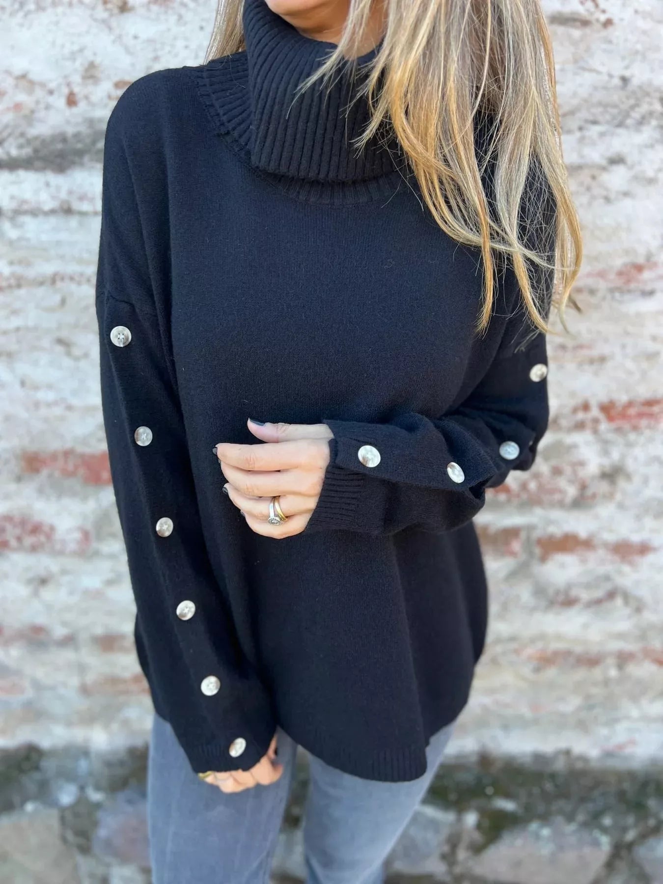 Woman wearing a black ribbed turtleneck sweater with decorative button sleeves and jeans.