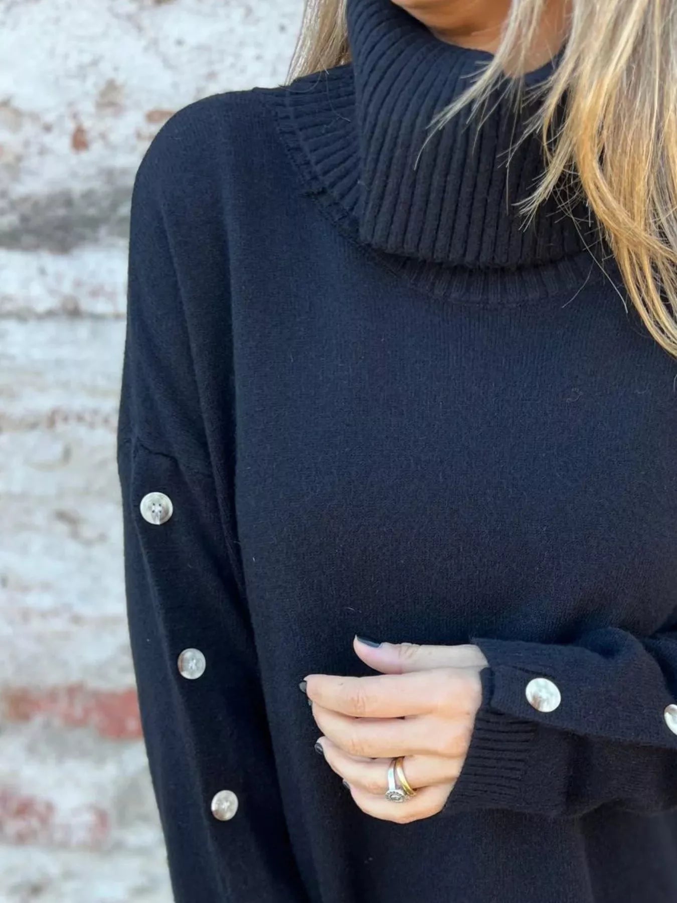 Woman wearing a black ribbed turtleneck sweater with decorative button sleeves and jeans.