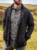 Men's black chunky knit shawl collar cardigan with button-front closure, ribbed texture, and front pockets, layered over a gray button-down shirt.