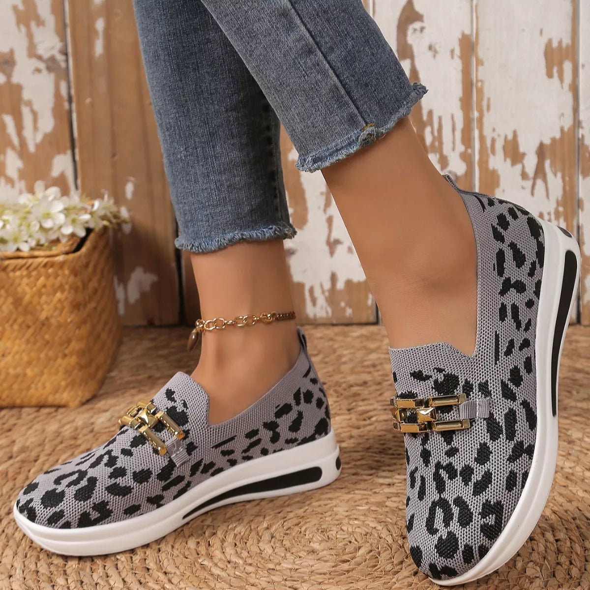 Leopard print slip-on shoes with gold accents worn by a person on a wooden floor.