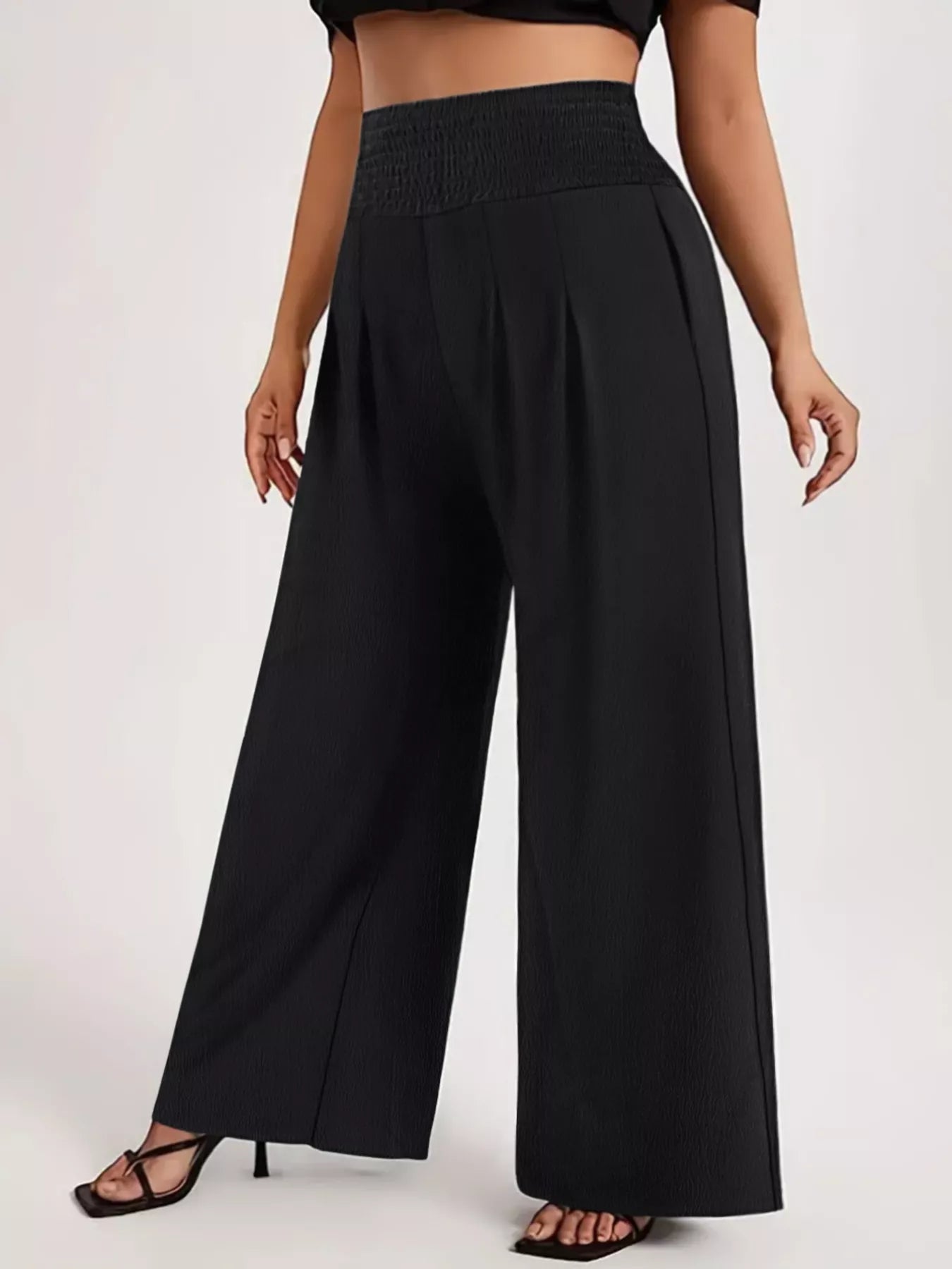 Model wearing black plus-size high-waisted wide-leg pants with a smocked waistband and side pockets.