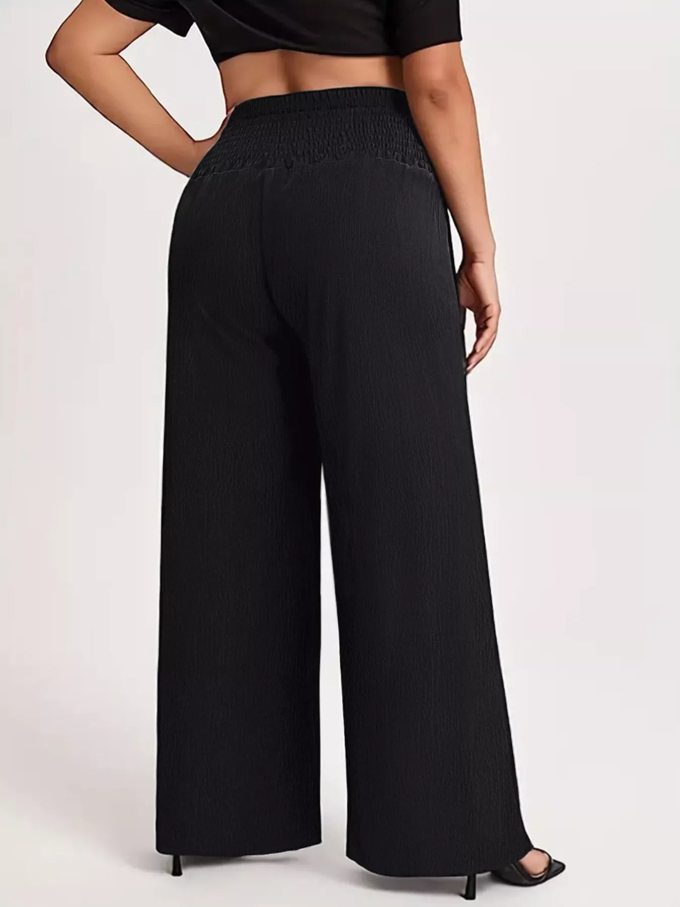 Model wearing black plus-size high-waisted wide-leg pants with a smocked waistband and side pockets.