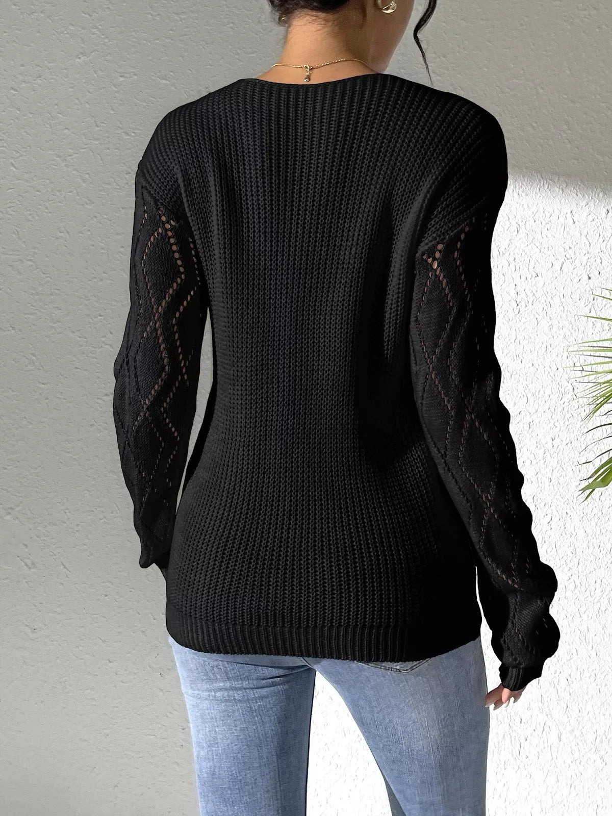 Woman wearing a black V-neck hollow knit sweater with contrasting neckline, openwork long sleeves, and blue jeans.