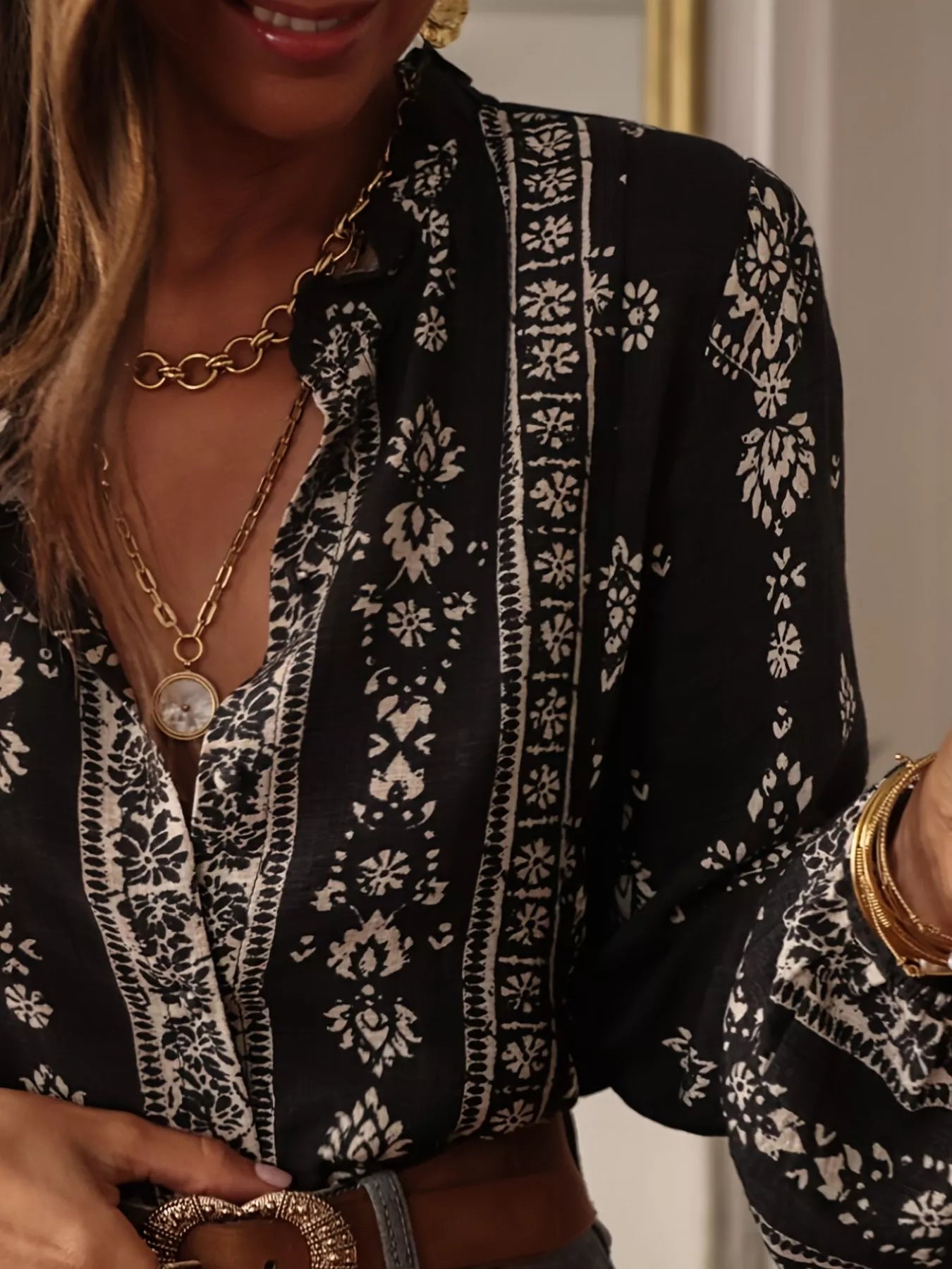 Woman wearing a beige and black vintage print long-sleeve blouse with dark denim jeans, a brown belt, and statement jewelry.