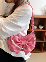 Person holding a pink handbag with a bow and pearl chain in front of a wooden shelf.