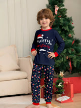 Boy wearing matching navy Merry Christmas pajama sets with red cuffs and holiday-print pants beside a decorated Christmas tree.