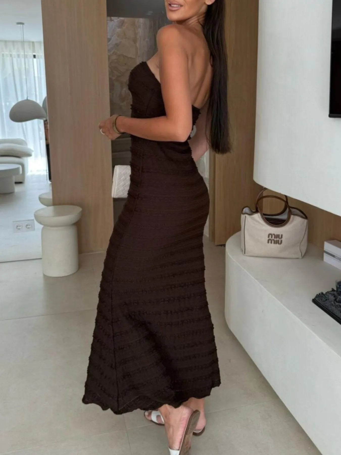 Woman wearing a brown sweetheart neck tube maxi dress with ruched detailing and a subtle cutout, styled with an ivory clutch for an elegant evening look.