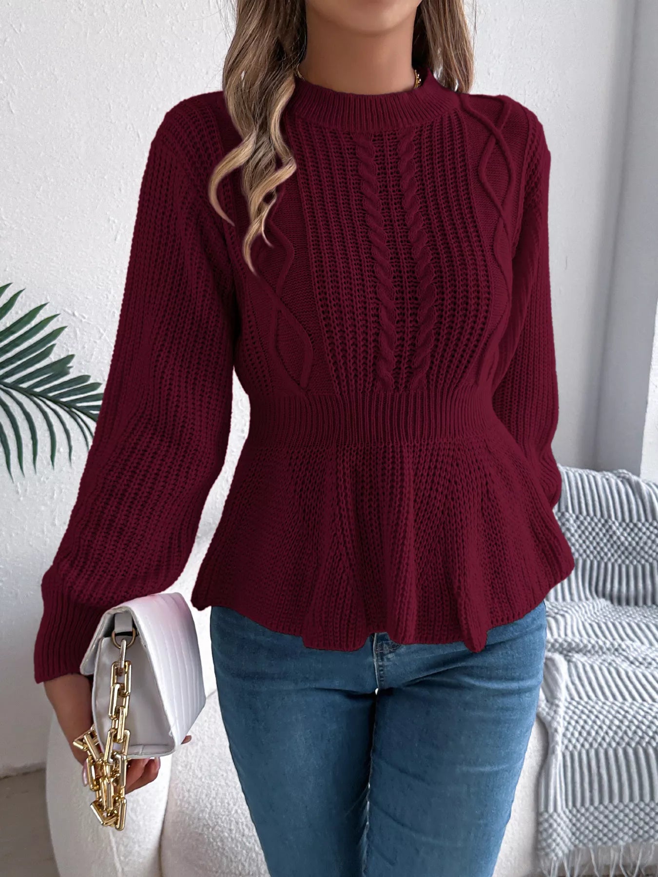 Woman wearing a burgundy cable knit round neck peplum sweater with long sleeves and blue jeans.