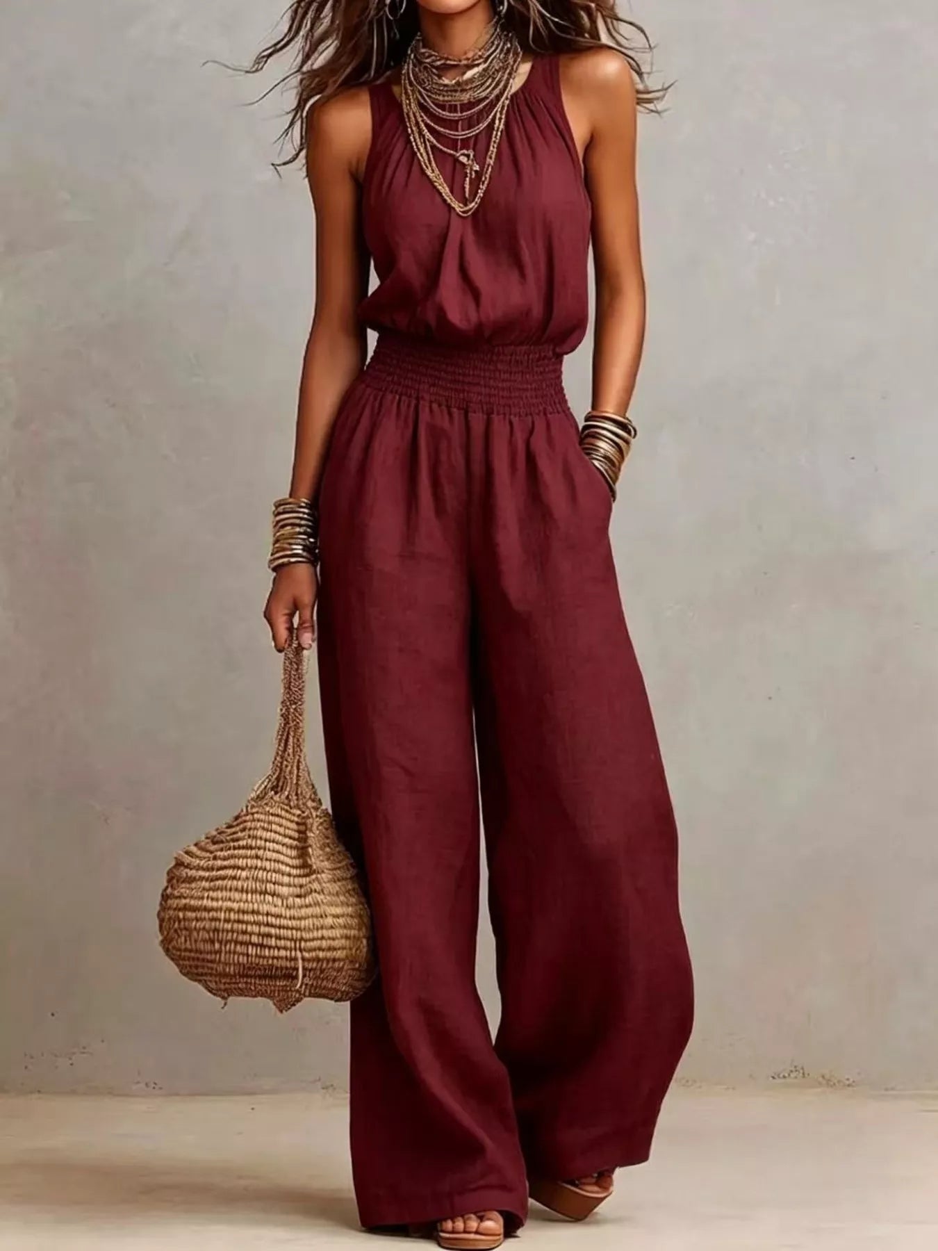 Woman wearing a burgundy halter-neck wide-leg jumpsuit with a smocked waist, layered gold jewelry, and bracelets, styled for an elegant evening look.