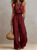 Woman wearing a burgundy halter-neck wide-leg jumpsuit with a smocked waist, layered gold jewelry, and bracelets, styled for an elegant evening look.