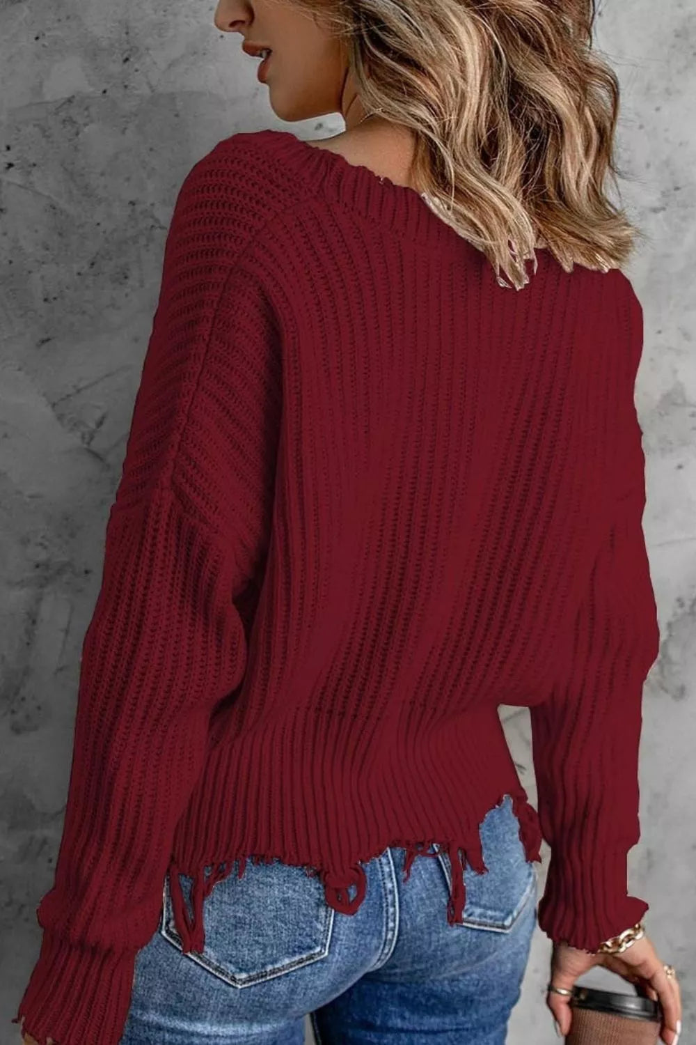 Woman wearing a burgundy ribbed V-neck sweater with distressed raw hem and blue jeans.