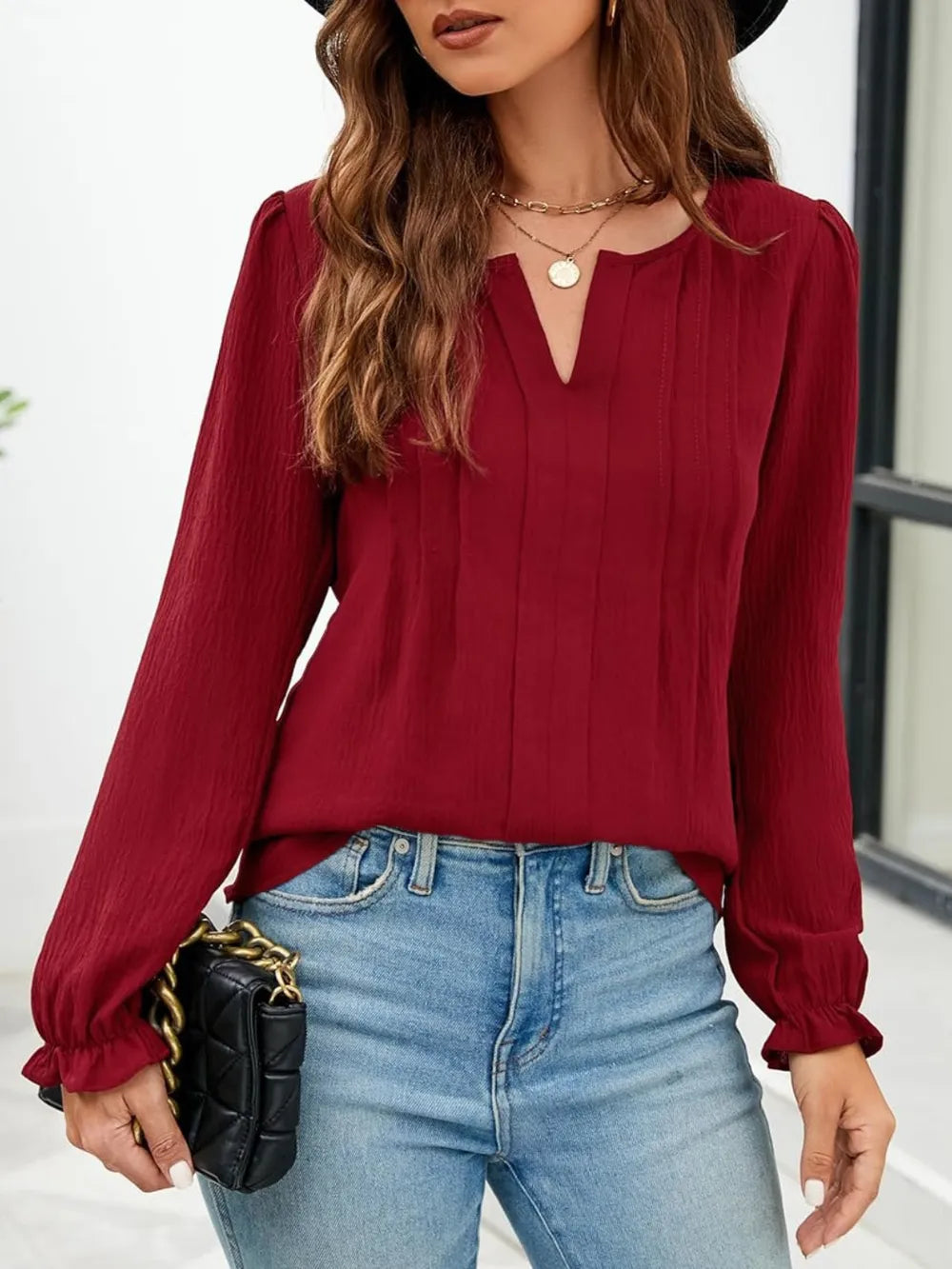 Woman wearing a burgundy notched neck long sleeve blouse with flounce cuffs styled with light-wash jeans and a black handbag.