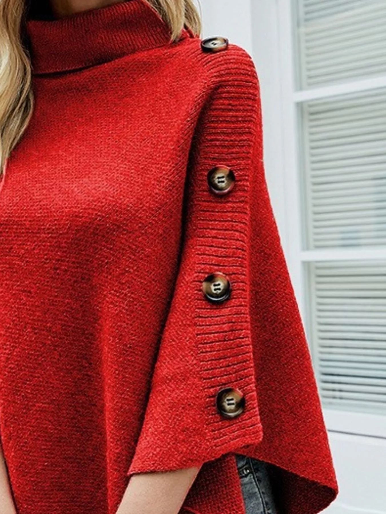 Women's red knit turtleneck poncho pullover with decorative buttons along the sleeves and pointed asymmetrical hem, styled with light-wash jeans.