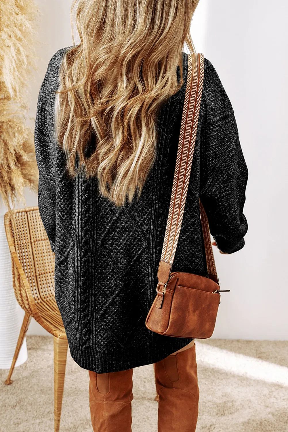 Woman wearing a black cable knit long sleeve sweater dress styled with knee-high brown boots and layered gold necklaces.