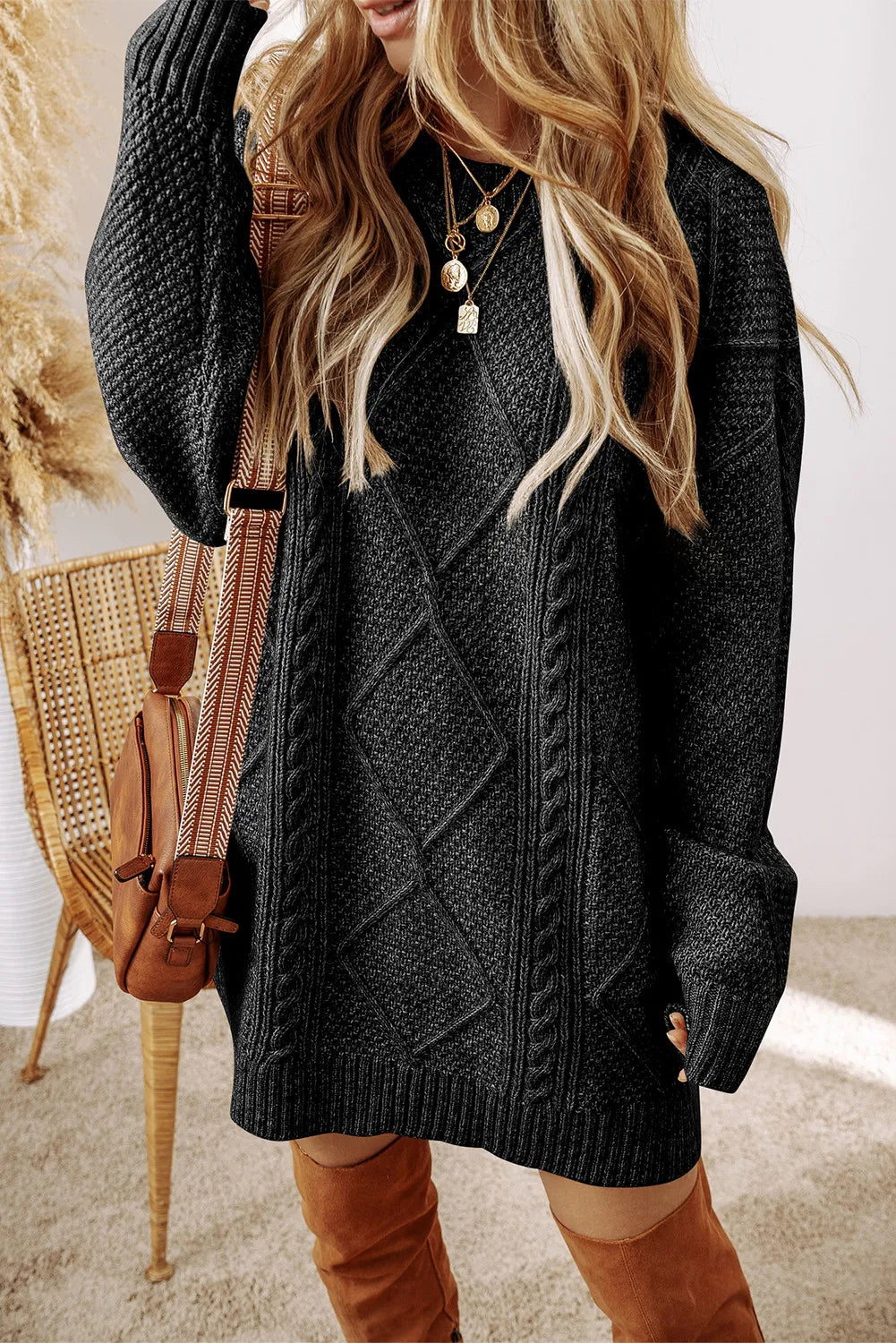 Woman wearing a black cable knit long sleeve sweater dress styled with knee-high brown boots and layered gold necklaces.