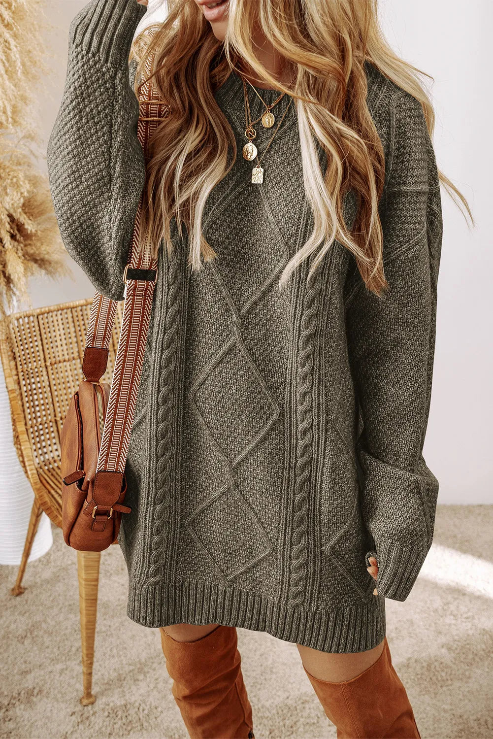Woman wearing a sage green cable knit long sleeve sweater dress styled with knee-high brown boots and layered gold necklaces.