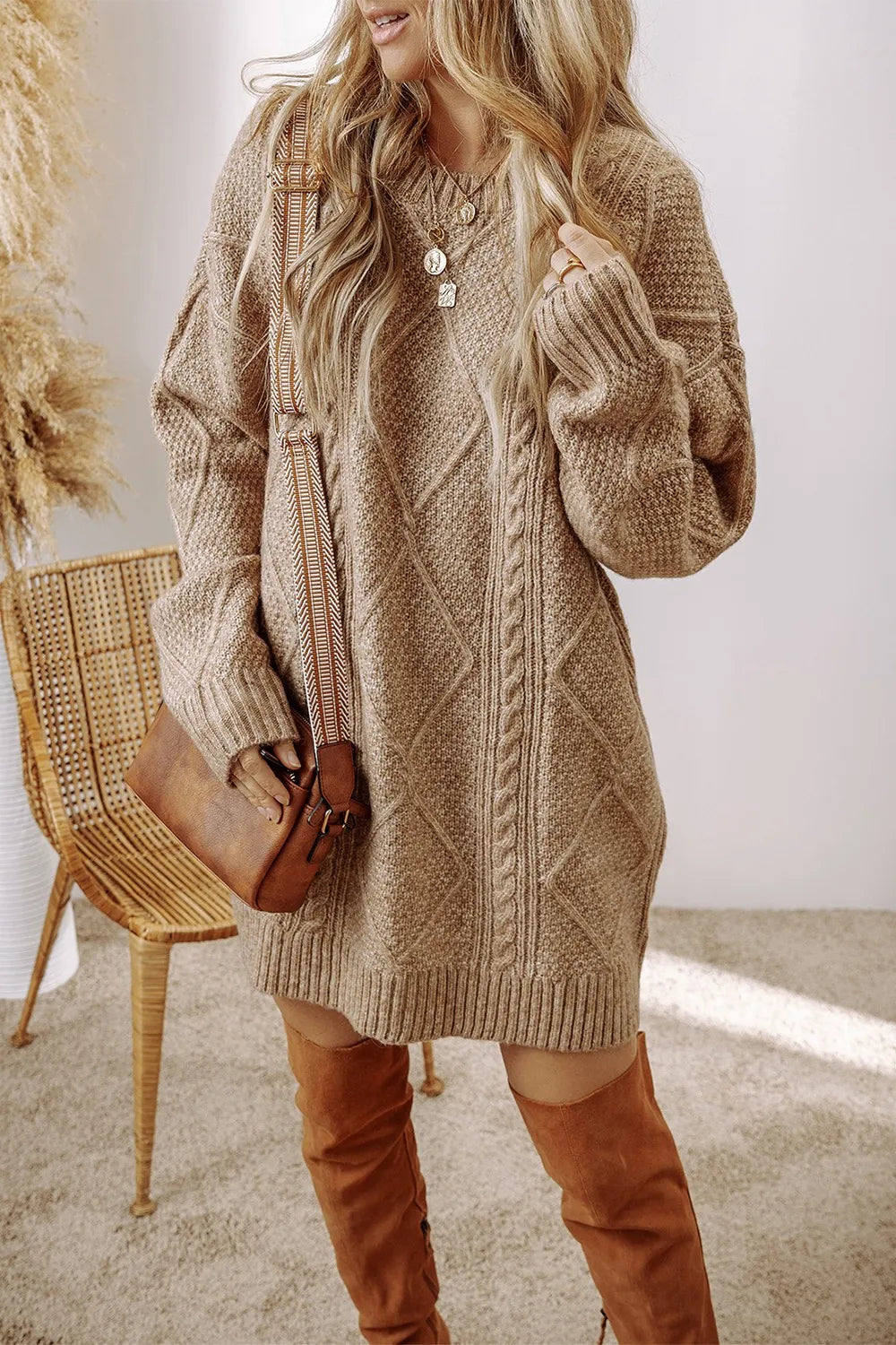Woman wearing a tan cable knit long sleeve sweater dress styled with knee-high brown boots and layered gold necklaces.