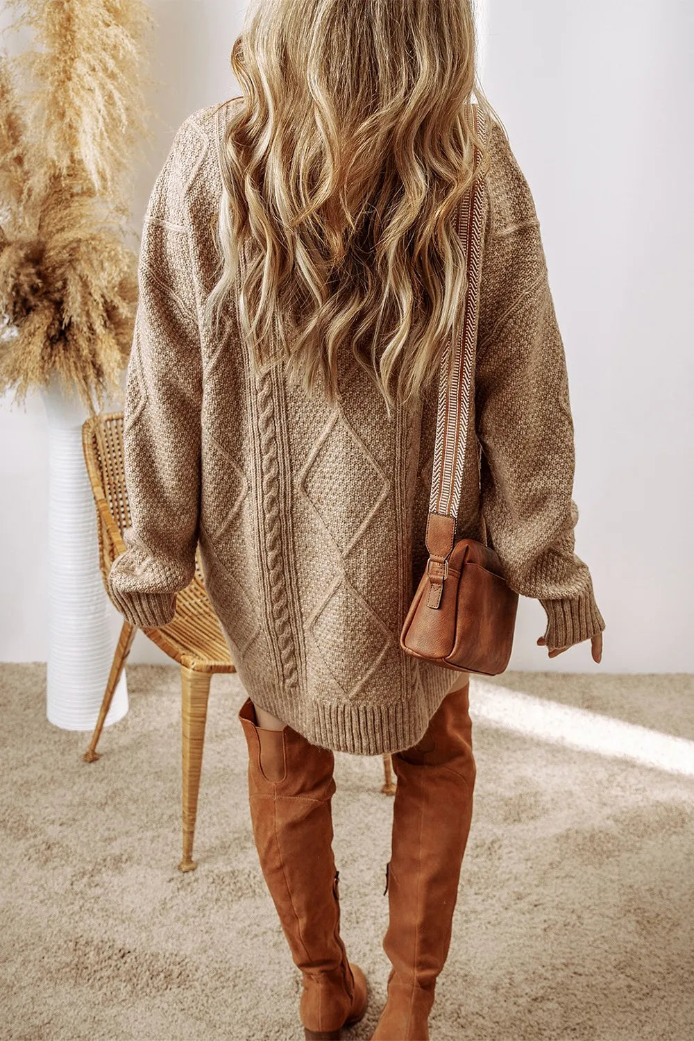 Woman wearing a tan cable knit long sleeve sweater dress styled with knee-high brown boots and layered gold necklaces.