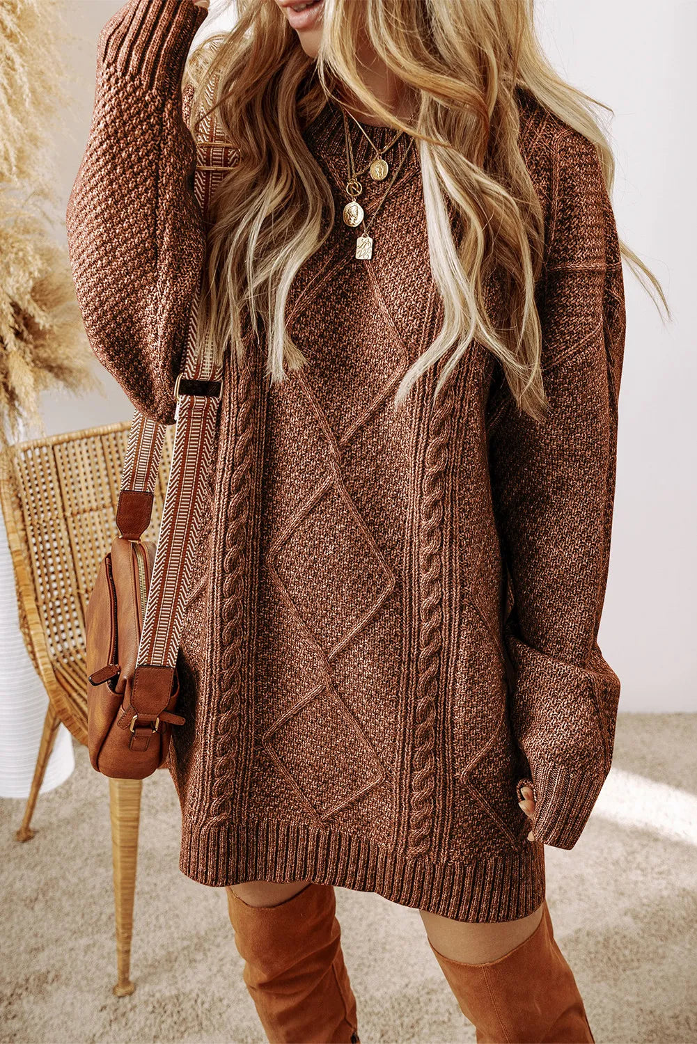 Woman wearing a taupe cable knit long sleeve sweater dress styled with knee-high brown boots and layered gold necklaces.