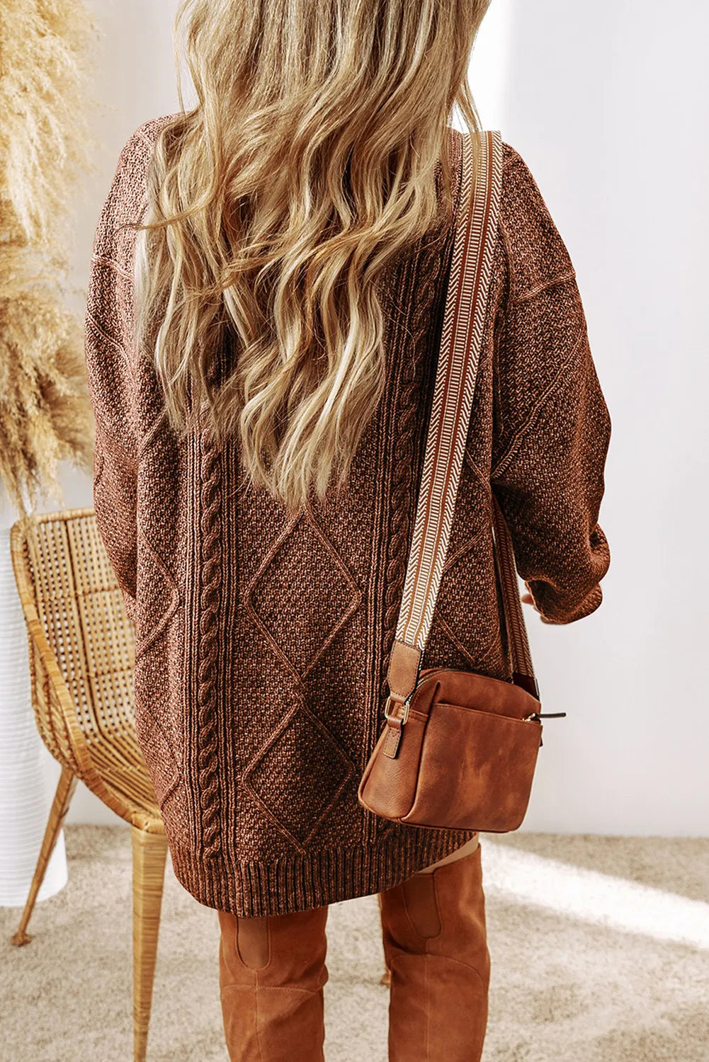 Woman wearing a taupe cable knit long sleeve sweater dress styled with knee-high brown boots and layered gold necklaces.