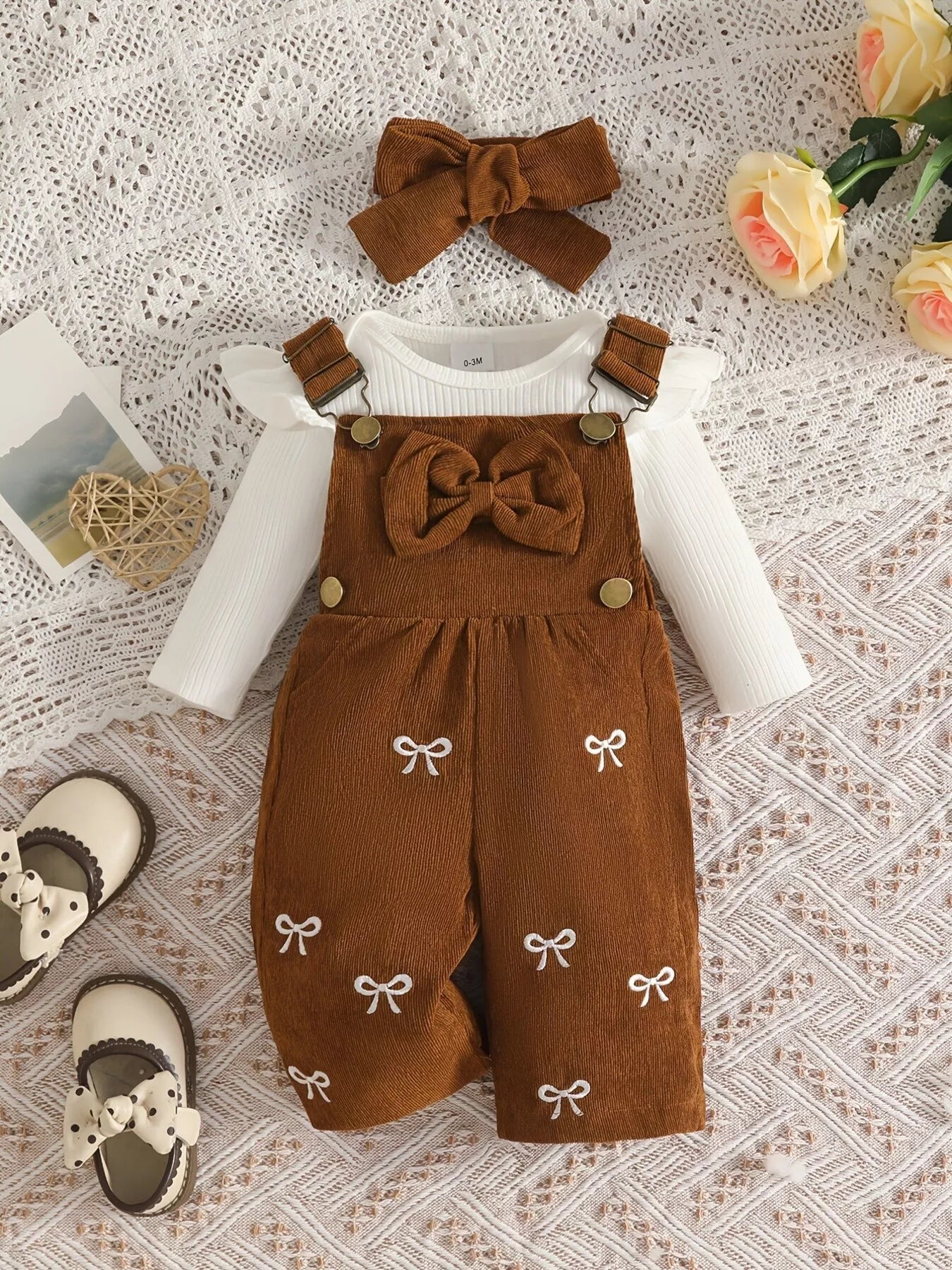 Baby girl three-piece outfit featuring a cream ribbed long-sleeve bodysuit with ruffled shoulders, caramel color bow-embroidered overalls and matching caramel color bow headband.