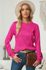 Woman wearing a cerise textured round neck long sleeve blouse with dark-wash jeans, brown clutch and cream hat.