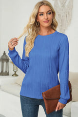 Woman wearing a cerulean textured round neck long sleeve blouse with dark-wash jeans and brown clutch.