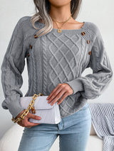 Woman wearing a charcoal cable-knit sweater with decorative buttons, paired with light-wash jeans, layered gold necklaces, and a white handbag. Perfect for cozy fall and winter outfits.