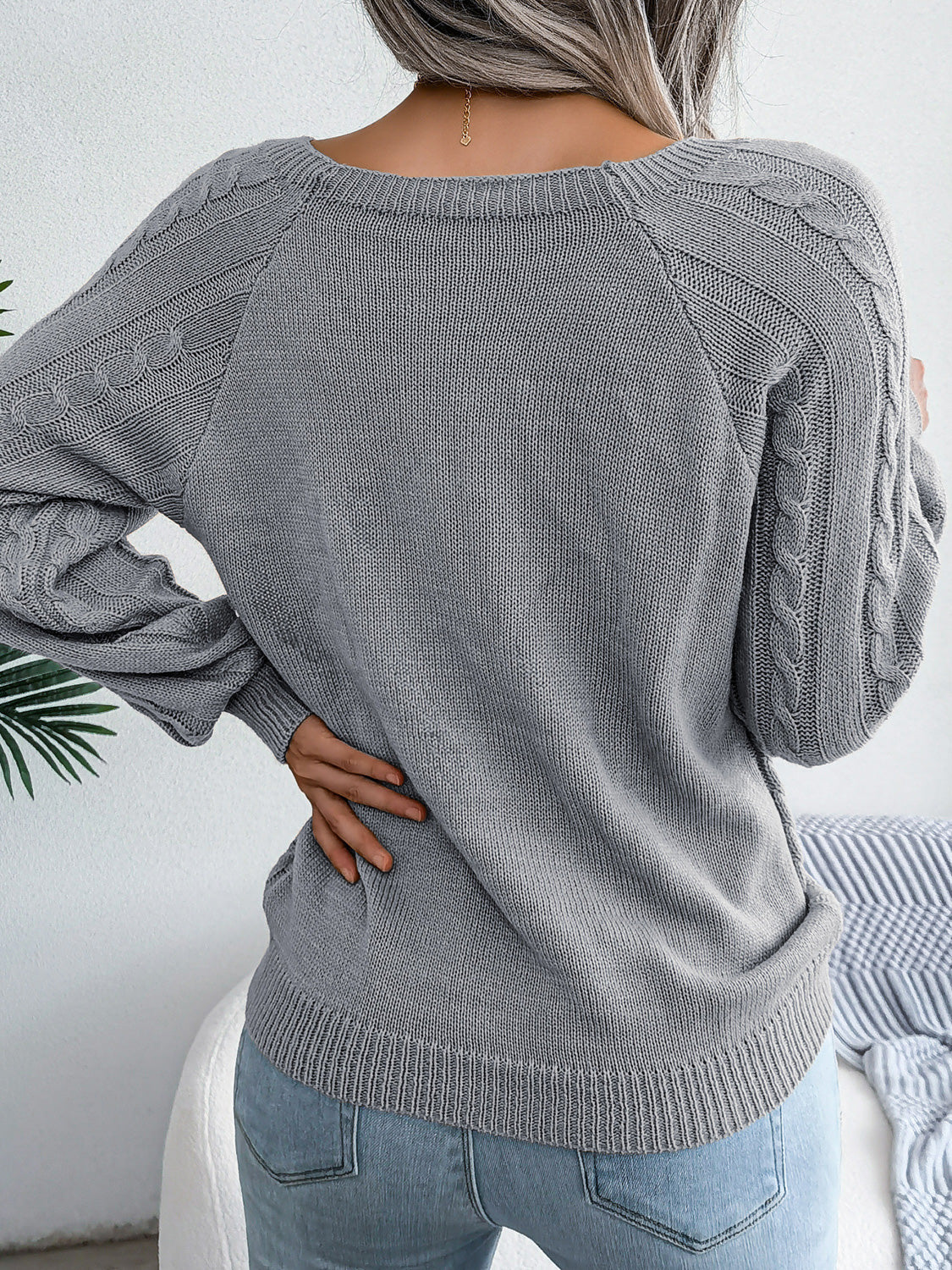 Woman wearing a charcoal cable-knit sweater with decorative buttons, paired with light-wash jeans, layered gold necklaces, and a white handbag. Perfect for cozy fall and winter outfits.