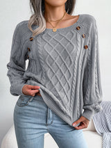 Woman wearing a charcoal cable-knit sweater with decorative buttons, paired with light-wash jeans, layered gold necklaces, and a white handbag. Perfect for cozy fall and winter outfits.