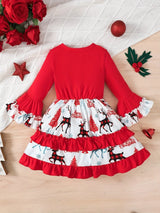 Little girl wearing a red long-sleeve Christmas dress with reindeer print, layered ruffle skirt and festive patterned sleeve cuffs beside a decorated Christmas tree.