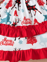 A red long-sleeve Christmas dress with reindeer print, layered ruffle skirt and festive patterned sleeve cuffs beside a decorated Christmas tree.
